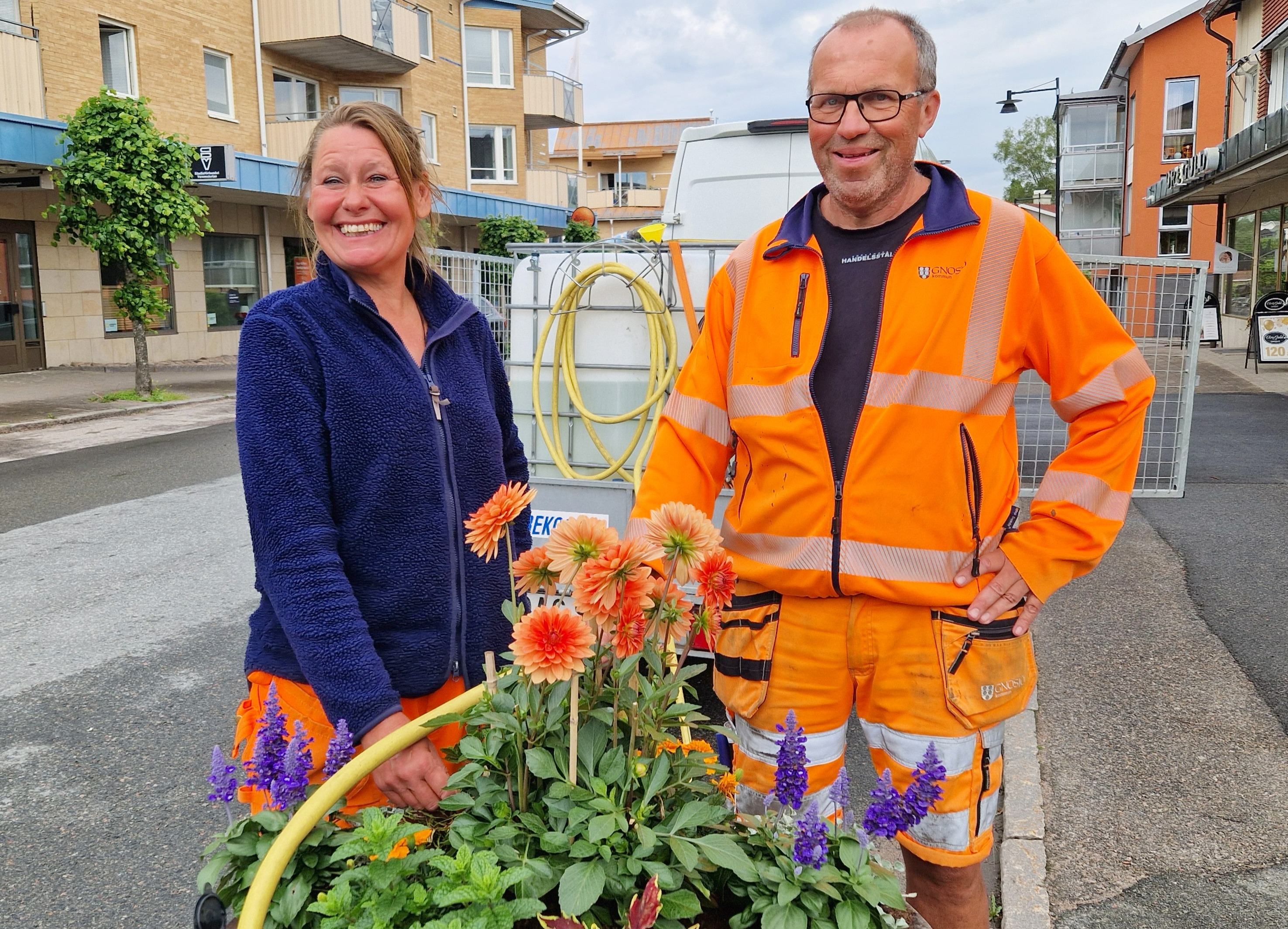 En kvinna och en man står vid en står blomkrika och vattnar.