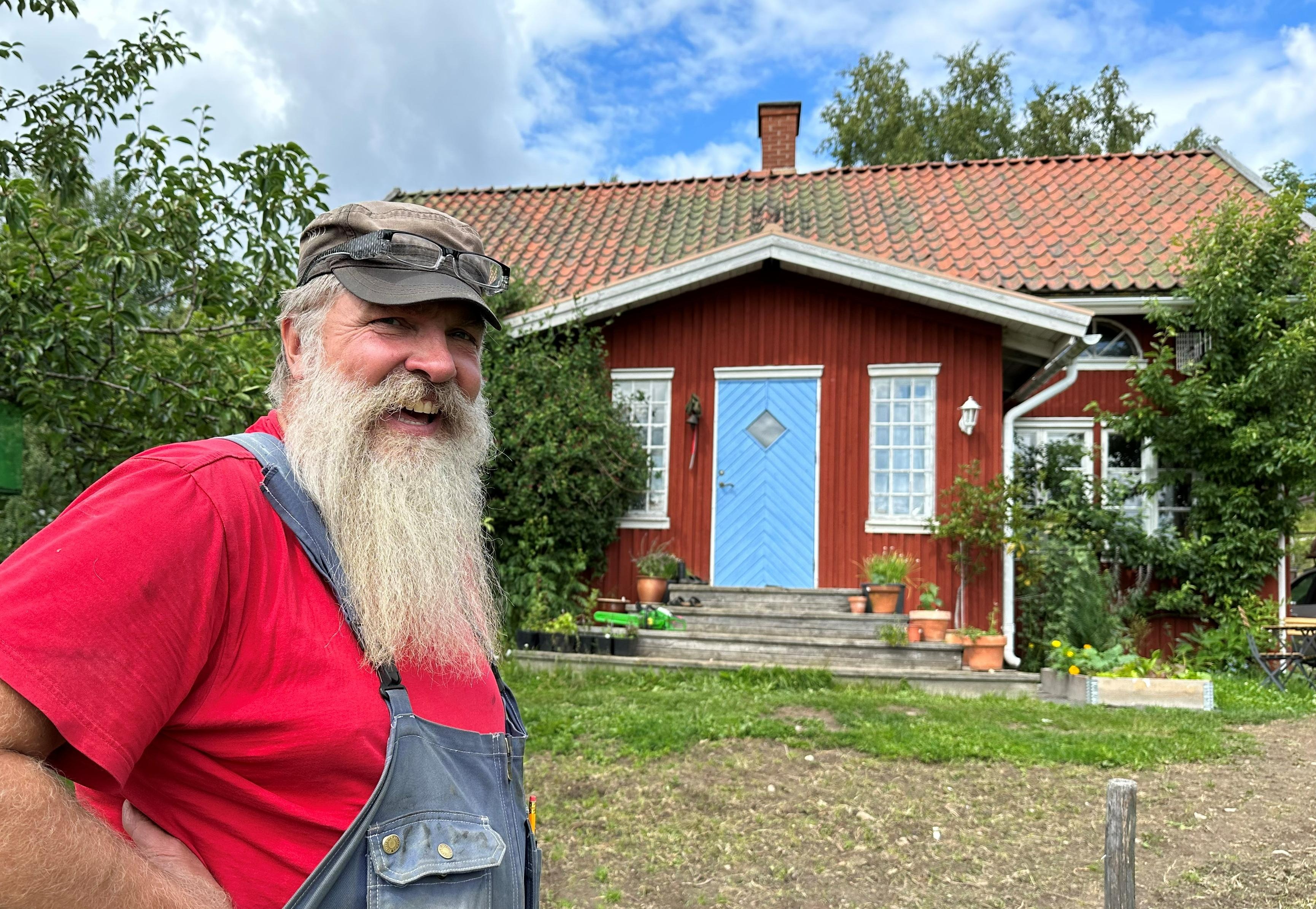 En glad man med stort skägg står framför ett rött hus med blå dörr.
