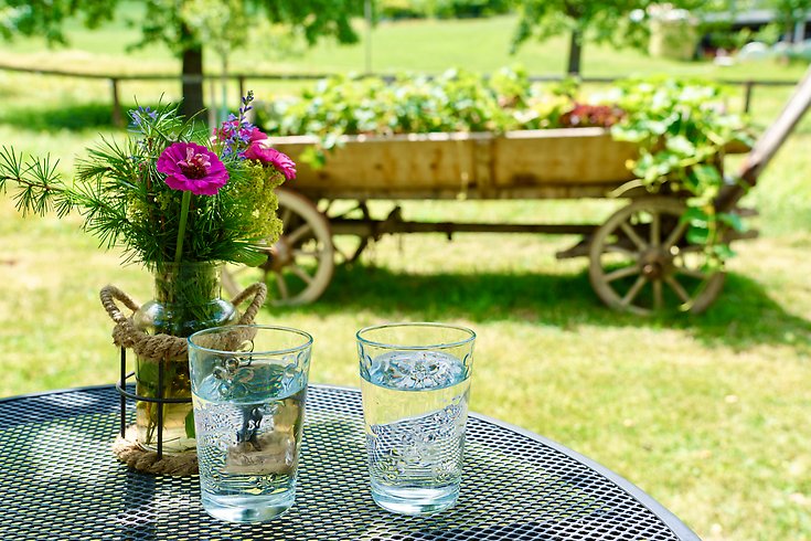 Två vattenglas står på ett bord framför en gammal skrinda. På bordet står också en bukett med lila blommor.