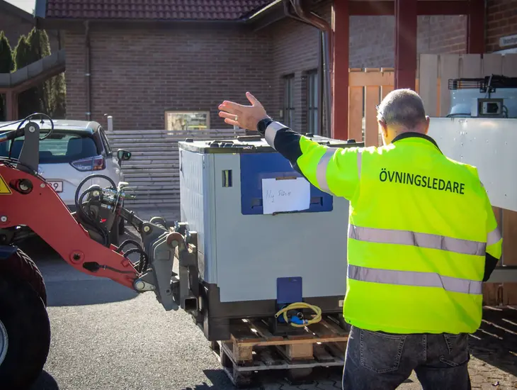 En man i varselväst med texten "övningsledare" instruerar en traktor med en vattentank.