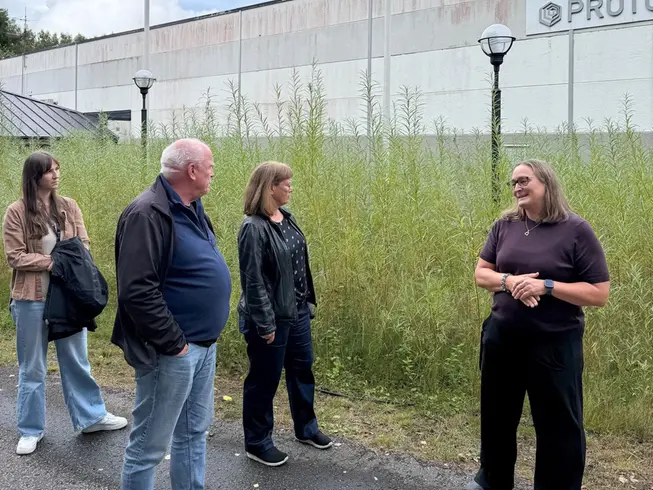 En grupp människor utanför en industribyggnad, ett fält med växtlighet i bakgrunden.
