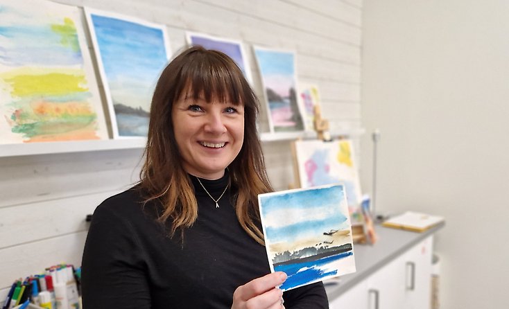 En glad kvinna håller upp en akvarell med ett landskap med tydlig horisont på.