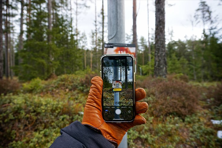 En hand med brun handske håller upp en mobil som scannar en QR-kod för att läsa frågan.